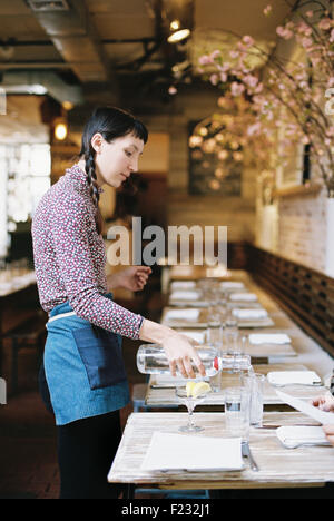 Eine Frau trägt eine Schürze gießt Wasser in Gläser am Tisch in einem Restaurant. Stockfoto