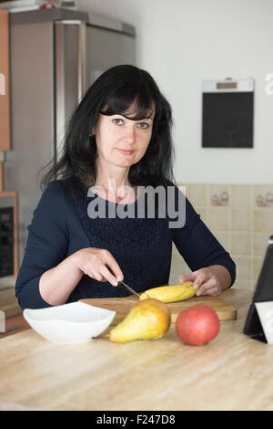 Lächelnde Frau schneiden Banane in Küche auf Schneidebrett zu einem Fruchtcocktail Stockfoto