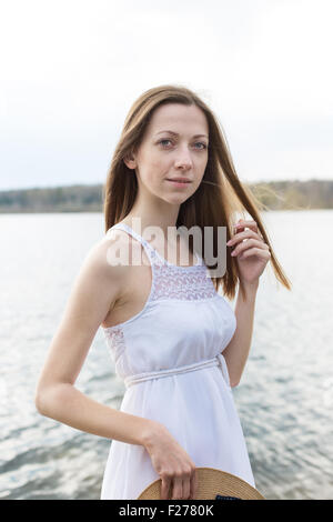 Sommersprossige glückliches Mädchen im weißen Kleid Stroh Hut in den Händen hält und Blick in die Kamera bei windigem Wetter Stockfoto