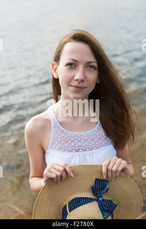 Sommersprossige glückliches Mädchen im weißen Kleid Stroh Hut in ihren Händen hält und Blick in die Kamera bei windigem Wetter Stockfoto