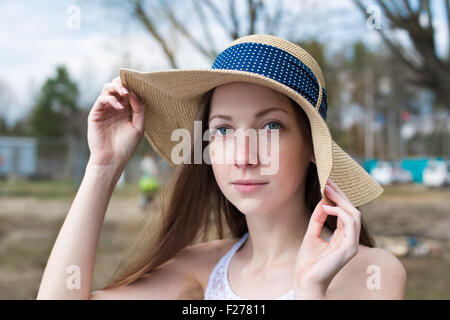 Sommersprossige glückliches Mädchen im weißen Kleid hält Stroh Hut und Blick in die Kamera bei windigem Wetter Stockfoto