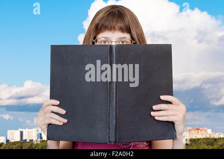 Mädchen mit Brille blickt auf große Buch mit Blindabdeckung und weiße Wolke über der Stadt auf Hintergrund Stockfoto