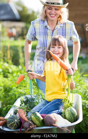 Kind mit Spaß in einer Schubkarre schieben von Mutter im heimischen Garten auf warmen, sonnigen Sommer. Stockfoto