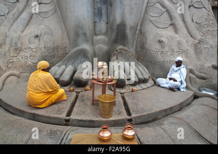 Jaïn Pilger (der Mann in Orange gekleidet) und ein Priester sitzen an den Füßen der Statue des Gomateshwara (Indien) Stockfoto