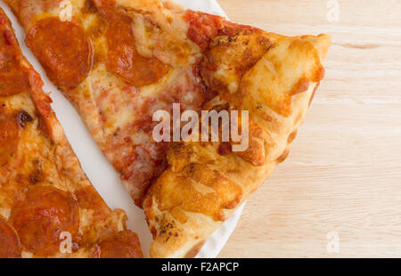 Draufsicht enger auf zwei Scheiben frisch gebackene Peperoni-Pizza auf einem weißen Papierteller oben auf einer hölzernen Tischplatte Stockfoto