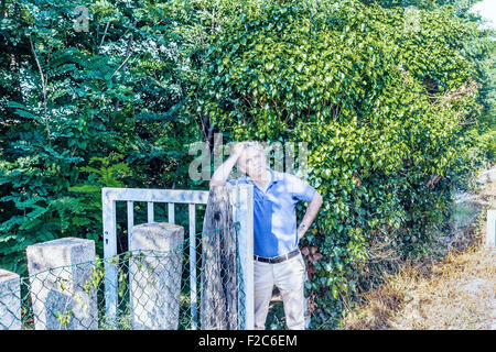 Kaukasischer Mann mittleren Alters, elegant und sportlich kleiden einen blauen Polo hat einen nachdenklichen Blick während Kopf und lehnte sich gegen ein Stahltor in der italienischen Landschaft im Sommer Stockfoto