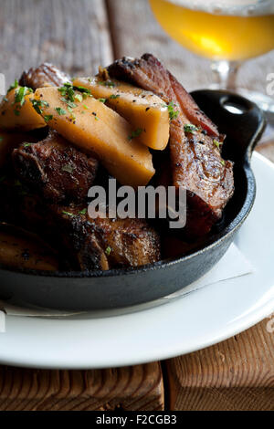 Langsam gebratene Schweinerippchen mit karamellisierten Äpfeln in schwarzem Gusseisen Pfanne auf Tisch mit Bier Stockfoto