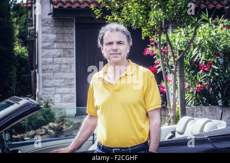 Edel 40 Jahre alten Sportler mit drei-Tage-Bart und Salz und Pfeffer Haar trägt ein gelbes Poloshirt, während er an der Tür eines dunklen braunen Cabriolet Autos in Wohngegend sitzt Stockfoto