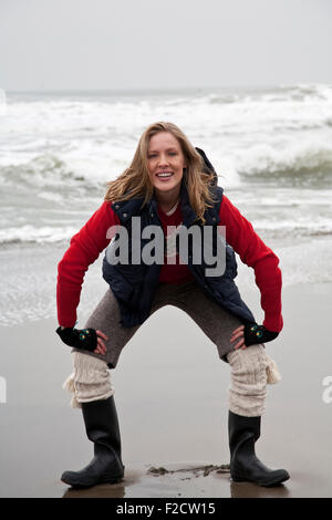 Junge, blonde Frau steht vor Kamera, hocken im Winter am Strand vor Ozean Stockfoto