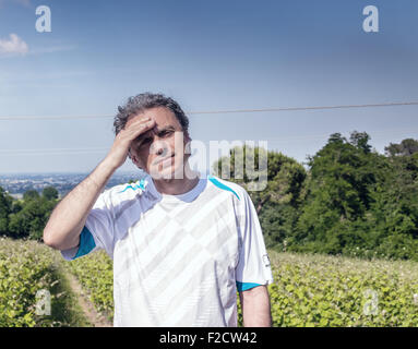 Gut aussehend 40 Jahre alter Mann mit Salz Pfeffer Haar gekleidet mit Sport-Shirt hält seinen Kopf in den Feldern der italienischen Landschaft: er scheint Kopfschmerzen haben Stockfoto