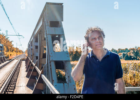 Kaukasische 40 Jahre alter Mann, elegant und sportlich kleiden ein blaues Polo-Shirt, spricht auf dem Handy neben Bahngleisen in der italienischen Landschaft im Sommer Stockfoto