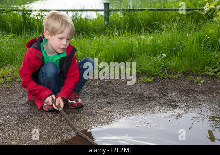 blonde junge spielt mit Stock in eine Pfütze Stockfoto