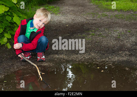 blonde junge spielt mit Stock in eine Pfütze Stockfoto