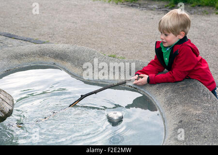 blonde junge spielt mit Stick bei der Fontäne Stockfoto