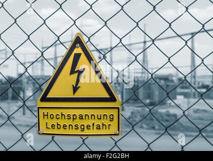 gelbe Deutsche Hochspannung Warnschild auf einem Gitter Drahtzaun vor einem blau getönten Umspannwerk in Süddeutschland Stockfoto