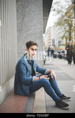 schönen jungen alternative dunklen Mann sitzt auf dem Boden mit Smartphone suchen unter Ausschluss der Öffentlichkeit mit zentralen Labret und Nasenloch piercing - Technik, soziales Netzwerk, Kommunikations-Konzept Stockfoto