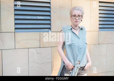 Halblängen eine junge hübsche kaukasischen blonde italienische Designer posiert Hände an eine Wand gelehnt, in Tasche, suchen in der Kamera - Tragens Azure Kleid und gesprenkelten Gläser - Jugend, nachdenklich-Konzept Stockfoto