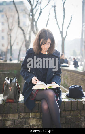 junge hübsche kaukasischen braune Haare Frau sitzt auf einer kleinen Mauer in der Stadt lesen Buch während Rauchen einer Zigarette mit Beine gekreuzt, auf der Suche nach unten - Kultur, relaxen Konzept Stockfoto