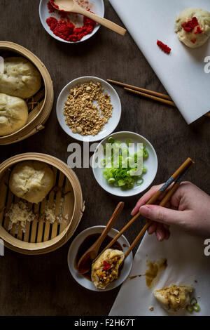 Zarte Frauenhand mit Stäbchen eintauchen gefüllte Knödel in Sauce auf hölzernen Tischplatte mit Bambus-Dampfer. Ansicht von oben Stockfoto