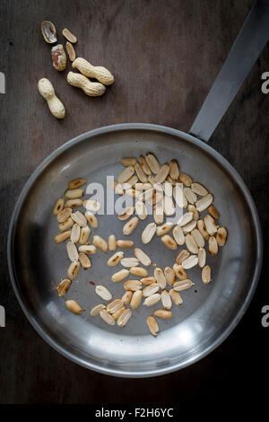Geröstete Erdnüsse in Stahl Pfanne auf hölzernen Tischplatte mit Erdnüsse in Schalen. Ansicht von oben Stockfoto