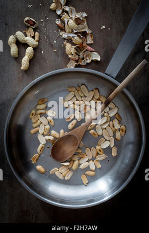 Geröstete Erdnüsse in Stahl Pfanne mit Holzspatel auf hölzernen Tischplatte mit geschälten und ganze Erdnüsse. Ansicht von oben Stockfoto