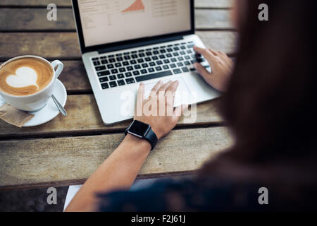 Frau Tippen auf der Tastatur eines Notebooks mit einer Kaffeetasse auf Holztisch. Frauen tragen eine Smartwatch im Café. Stockfoto