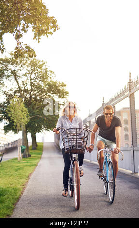 Romantisch zu zweit Radfahren auf einem Radweg in einem städtischen Park, Hand in Hand und lächelt in die Kamera an einem heißen Sommertag Stockfoto