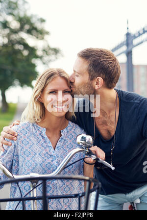 Liebevoller junger Mann seine Freundin auf der Stirn küssen, wie sie einen Sommertag an der frischen Luft auf dem Fahrrad genießen Stockfoto