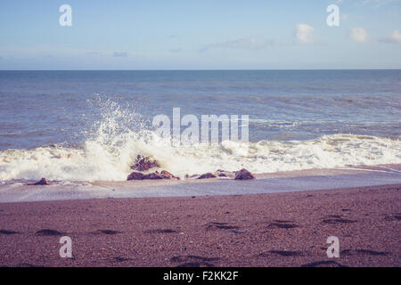 eine Welle stürzt auf einem kleinen Felsen an der Küste von einem feinen Kiesstrand, an dem Fußspuren im Sand gemacht wurden. Stockfoto