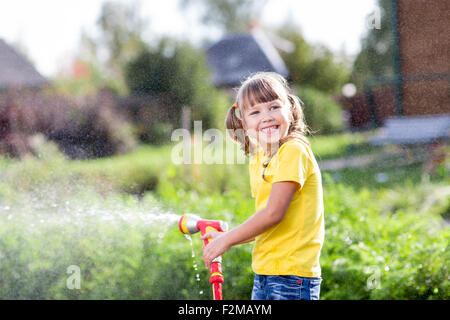 Fröhliches Kind Bewässerung von Pflanzen aus Schlauch Spray im Garten im Hinterhof des Hauses am sonnigen Sommer Stockfoto
