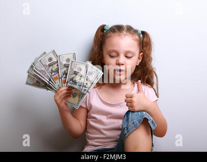 Glückliches Kind Mädchen halten Geld und denken, wie viele Dollar ich verdiente und zeigen Zeichen auf blauem Hintergrund Daumen Stockfoto