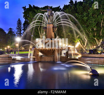 Vorderansicht des Sydney Hyde Park Wahrzeichen - Archibald Springbrunnen wie Sonnenuntergang mit Lichter Beleuchtung und unscharfen Wasserläufe Stockfoto