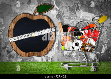 Warenkorb gefüllt mit Sportgeräten vor Steinmauer mit Apfel geformt Tafel Stockfoto