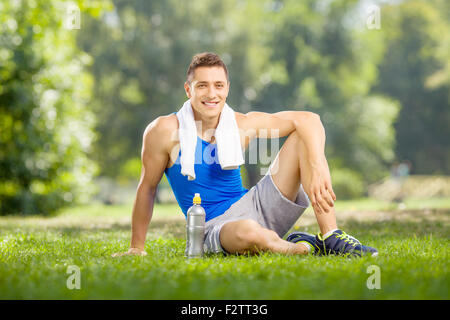 Junge Sportler in einem Park mit einer Flasche Wasser neben ihm zu sitzen und in die Kamera schaut Stockfoto