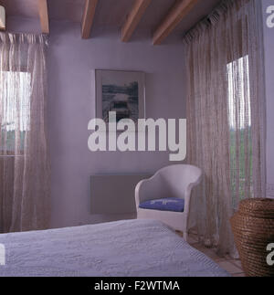 Weiße Gardinen am Fenster im Weißen Haus Schlafzimmer mit einem gemalten Lloyd Loom Stil Stuhl Stockfoto