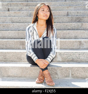 Porträt einer ziemlich asiatische Frau auf Treppen im Freien sitzen und auf der Suche nach etwas