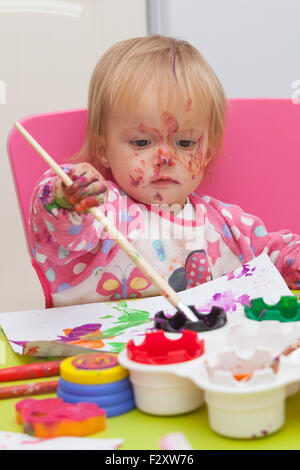 Ein 16 Monate alt, Blond Haare, Bürsten kaukasisch, Mädchen spielen mit Farben und die Farbe während des Sitzens im Inneren an einem kleinen Tisch. Stockfoto