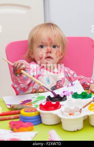 Ein 16 Monate alt, Blond Haare, Bürsten kaukasisch, Mädchen spielen mit Farben und die Farbe während des Sitzens im Inneren an einem kleinen Tisch. Stockfoto
