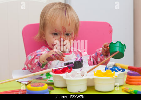Ein 16 Monate alt, Blond Haare, Bürsten kaukasisch, Mädchen spielen mit Farben und die Farbe während des Sitzens im Inneren an einem kleinen Tisch. Stockfoto