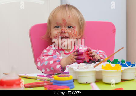 Ein 16 Monate alt, Blond Haare, Bürsten kaukasisch, Mädchen spielen mit Farben und die Farbe während des Sitzens im Inneren an einem kleinen Tisch. Stockfoto