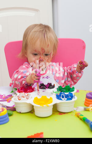 Ein 16 Monate alt, Blond Haare, Bürsten kaukasisch, Mädchen spielen mit Farben und die Farbe während des Sitzens im Inneren an einem kleinen Tisch. Stockfoto