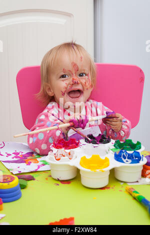 Ein 16 Monate alt, Blond Haare, Bürsten kaukasisch, Mädchen spielen mit Farben und die Farbe während des Sitzens im Inneren an einem kleinen Tisch. Stockfoto