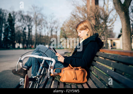 junge schöne Hipster sportliche blonde Frau sitzen auf einer Bank in einem Park mit Fahrrad und Tablet, auf der Suche nach unten den Bildschirm - Musik, sportliche Konzept Stockfoto