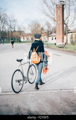 junge schöne Hipster sportliche blonde Frau zu Fuß in einem Stadtpark, halten Sie ein Fahrrad und ein Skate, trägt blaue Jeans, schwarzer Mantel, Hut und Rucksack - sportlich, Jugendkonzept - Ansicht von hinten Stockfoto