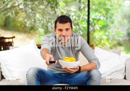 Hispanische Männchen tragen helle blauen Pullover plus Denim-Jeans sitzt im weißen Sofa hält Schüssel mit Pommes Frites und Fernbedienung Fernsehen mit Begeisterung Stockfoto