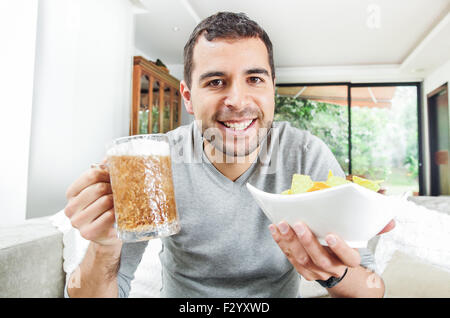 Closeup hispanische männlichen tragen helle blauen Pullover hält Bier und Schüssel mit Kartoffel-Chips vor Kamera, lustige Winkel Stockfoto