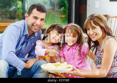 Hispanische Eltern mit zwei Töchtern aus einem Fach sitzt im Sofa und Lächeln und genießen einander Unternehmen Kartoffelchips Essen Stockfoto