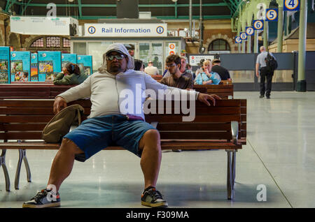 Ein übergewichtiger Mann sitzt auf einer Sitzbank mit gespreizten Beinen auf dem Landeisenbahnabschnitt des Hauptbahnhofs von Sydney in New South Wales, Australien Stockfoto