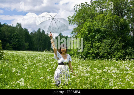 Junge Frau, brünett, Kaukasier, im weißen Kleid mit weißen Schirm auf einer Wiese in voller Blüte mit weißen Blüten. Stockfoto