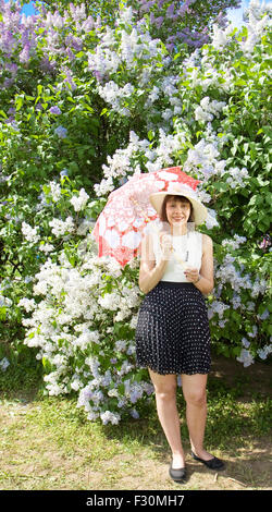 Hübsche Dame, brünett, europäischen, mit weißem Hut, weiße Bluse und schwarzen Rock mit weiß-roten Regenschirm in der Nähe von Flieder. Stockfoto